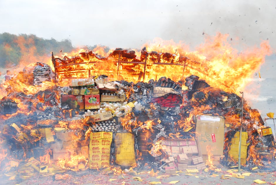 A large pile of household waste, including flattened cardboard boxes, plastic containers, and paper packaging, is engulfed in flames outdoors against a cloudy sky. The heap is situated on a grassy area, with some trash scattered on the ground nearby. Visible flames and smoke indicate an intense fire, with charred debris and partially burnt materials. In the background, faint structures or fences are partially obscured by smoke and heat. The image depicts a waste disposal or rubbish fire, illustrating the importance of proper bulky waste management and timely clearance. Man with Van Burnt Oak offers house removal and waste removal services which could help prevent such hazardous situations during home relocation or bulk waste disposal, ensuring compliance with local regulations in Barnet.