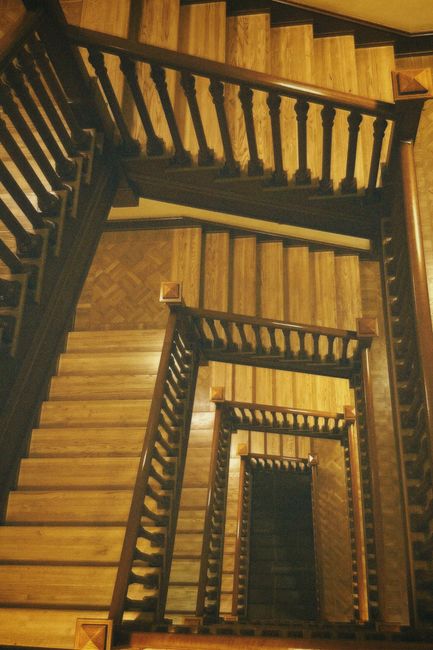 A view looking down a wooden staircase with multiple flights, carved wooden balustrades, and polished treads. The staircase is situated inside a property with warm-toned wooden flooring visible on the landings. The image captures the spiral arrangement of the stairs from an overhead perspective, showing the successive turns descending into the lower levels. The lighting is warm and highlights the natural wood grains of the staircase and floors. This setting may be part of a house undergoing relocation or furniture transport, with no moving equipment or objects visible in the image, focusing solely on the staircase structure. As part of a house removal service, Man with Van Burnt Oak handles such internal staircase navigation during home relocations, ensuring careful handling of furniture through narrow or complex staircases like this one.