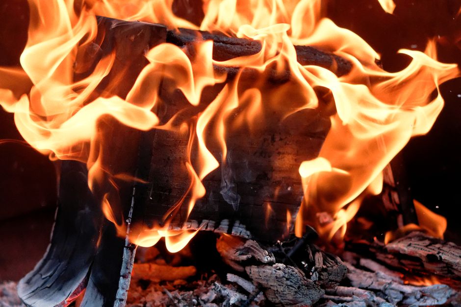 A close-up view of a wooden pallet with multiple sheets of paper stacked on top, set ablaze with bright orange and yellow flames. The fire illuminates the surrounding area against a dark background, h